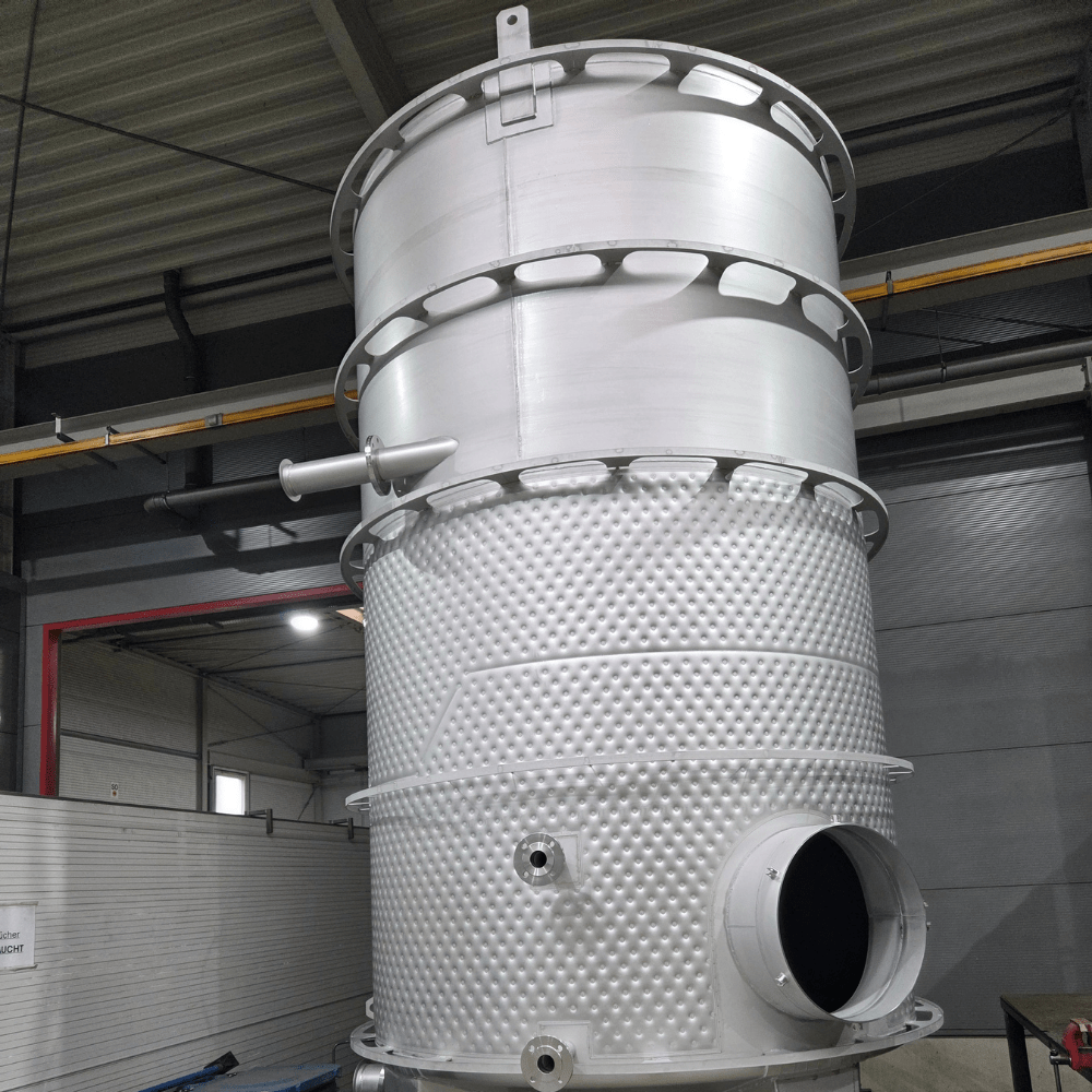Large stainless steel industrial vessel with a perforated lower section inside a factory setting, on a tall stand with circular rings at the top or middle part.