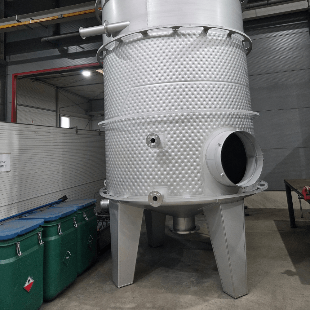 Industrial stainless steel processing vessel with a textured surface and large circular inlet, supported by sturdy legs in a warehouse setting.