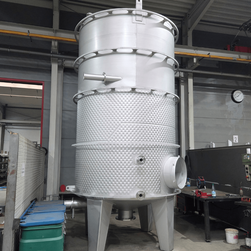 Large vertical metallic industrial vessel with a textured lower section and conical bottom supported on legs inside a factory, with side inlet/outlet pipe and top access features visible; background includes workshop equipment and a wall clock.