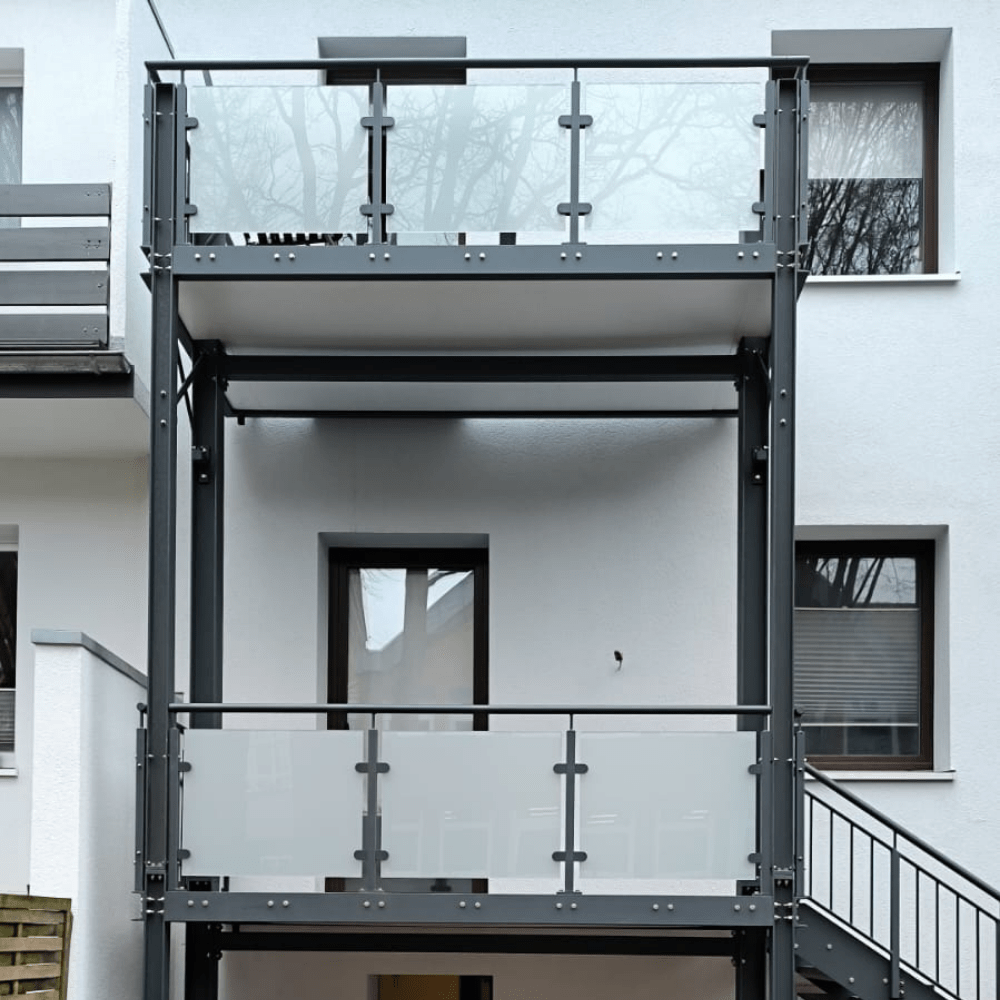 Modern balcony with a steel frame and frosted glass panels on a white apartment building, seen from the stairs corridor area.”,