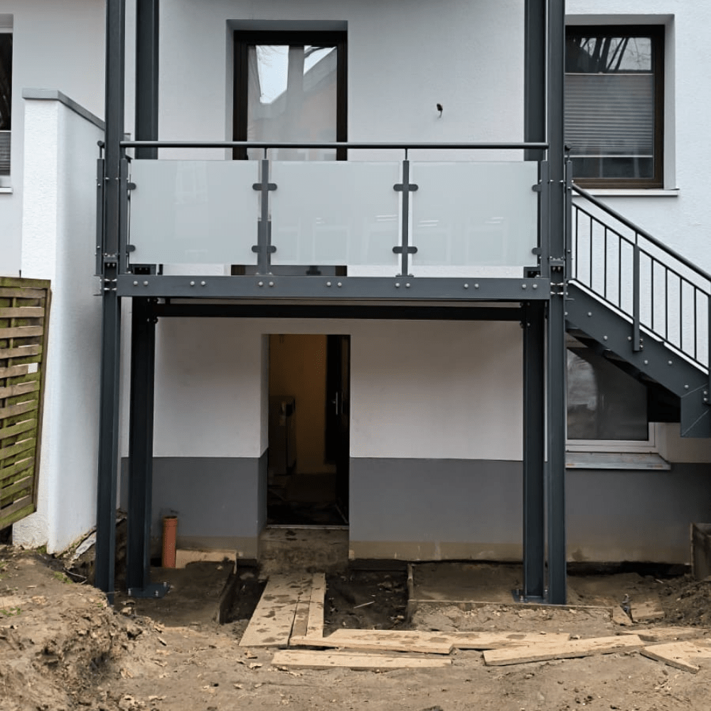 Open doorway to a basement under a second-floor balcony with glass panels and metal railings; construction debris and wooden planks on the dirt yard.